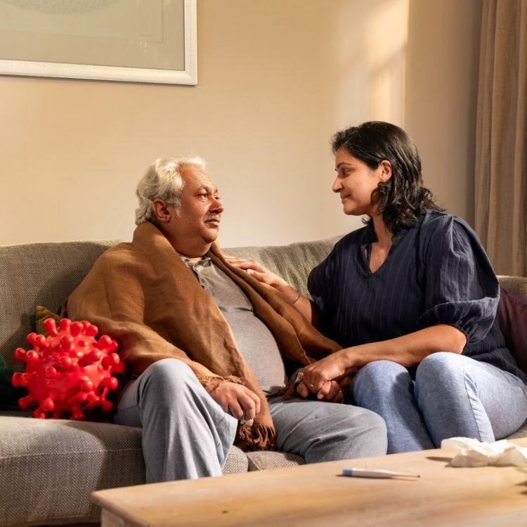 An older caucasian man sits on a couch wrapped in a blanket, being cared for by a younger woman. An englarged COVID-virus sits next to him.