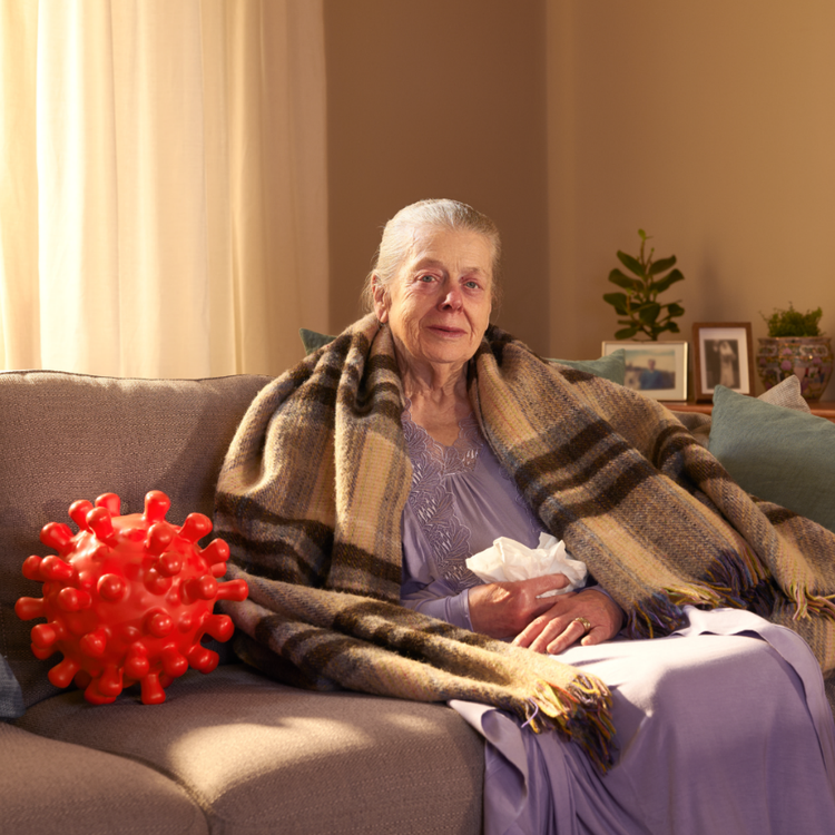 Sick elderly woman sitting on a couch, wrapped in a blanket, with tissues in her hand. An englarged COVID-virus sits next to her.