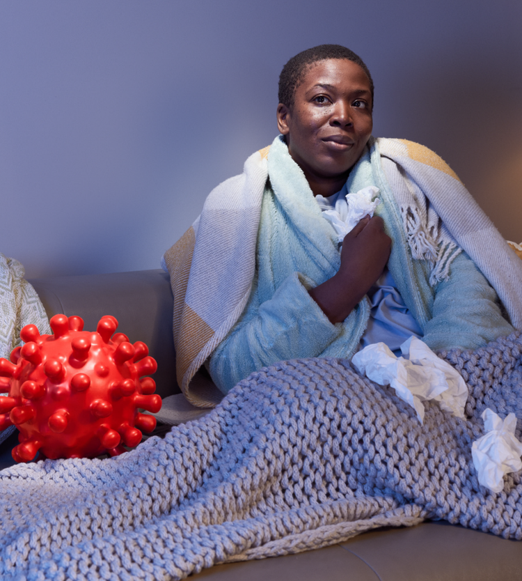 A sick African-American woman sits on the couch, wrapped in a blanket with an englarged COVID-virus next to her. A Know & Go-branded arrow sits in the bottom right corner, pointing to the right.