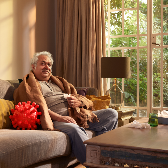 An older caucasian man sits on a couch wrapped in a blanket. An englarged COVID-virus sits next to him.