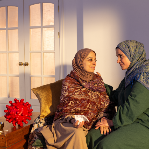 Two Middle Eastern women sit in a lviing room. One woman is sick, with tissues in hand, being comforted by the other woman. An englarged COVID-virus sits next to the sick woman.